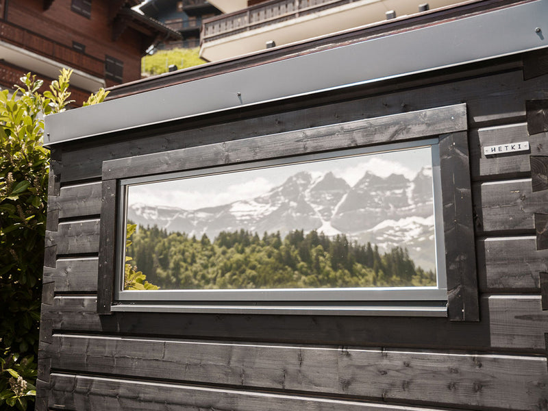 Dunkle Hetki Gartensauna mit großem Panoramafenster, in dem sich verschneite Berge spiegeln