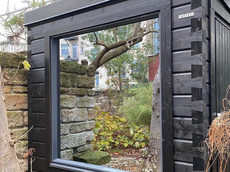 Dunkle Hetki Gartensauna mit großem Fenster in einem von Natursteinmauern umgebenen Stadtgarten