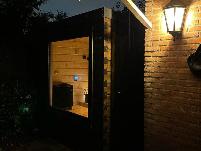 Beleuchtete schwarze Gartensauna neben einer Backsteinwand bei Nacht, Saunaofen im Fenster sichtbar