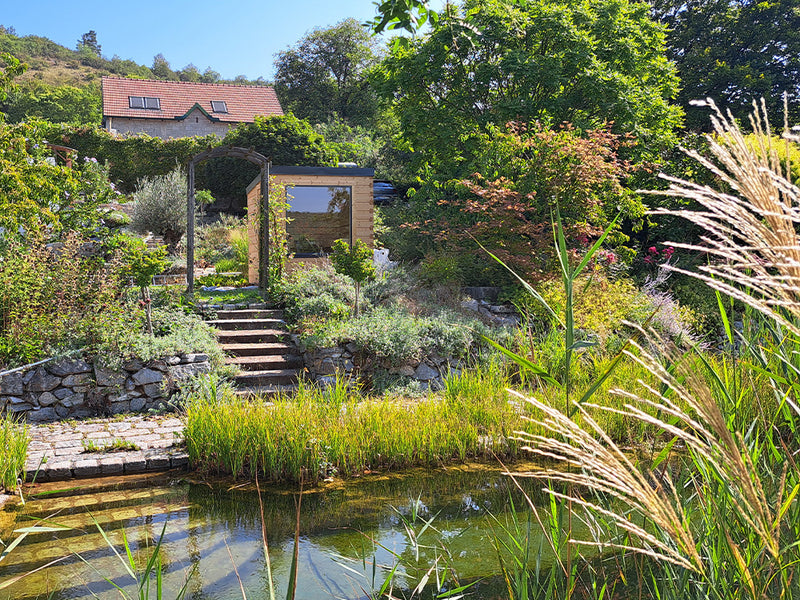 Kleine Gartensauna mit großem Fenster in einem üppig bepflanzten Garten am Teich