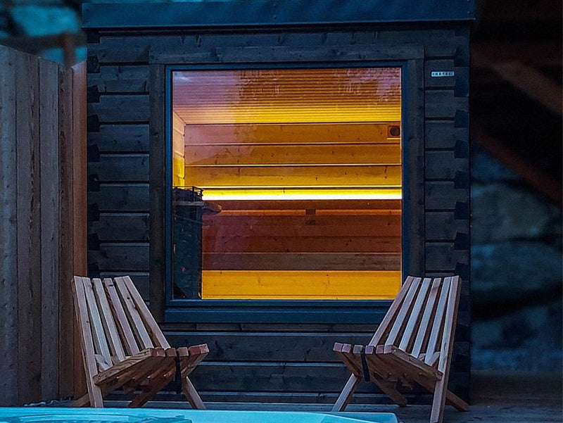 Beleuchtete dunkle Gartensauna bei Nacht mit großem Fenster und zwei Holzstühlen davor