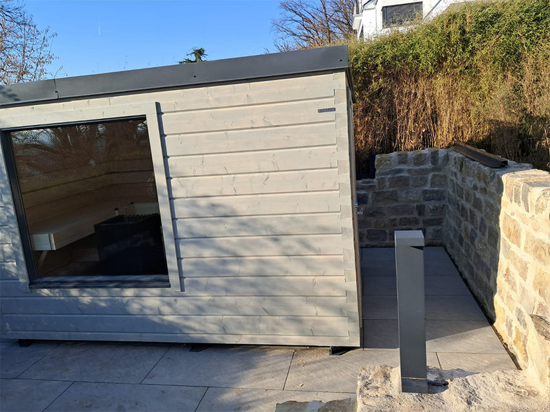 Graue Hetki Gartensauna mit großem Fenster zwischen Natursteinmauern