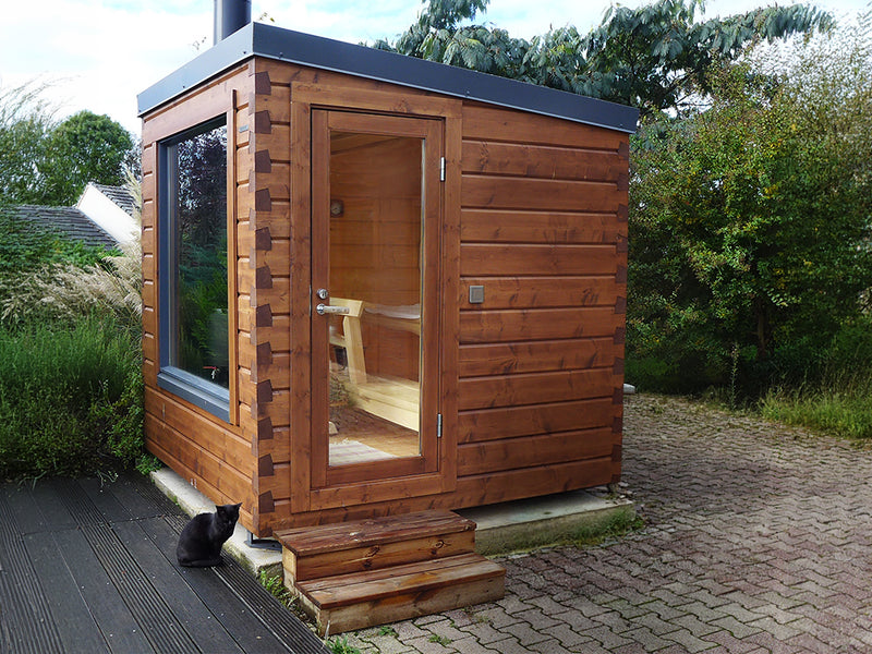 Braune Holz-Gartensauna mit Glastür, Seitenfenster und Stufen; eine schwarze Katze sitzt davor