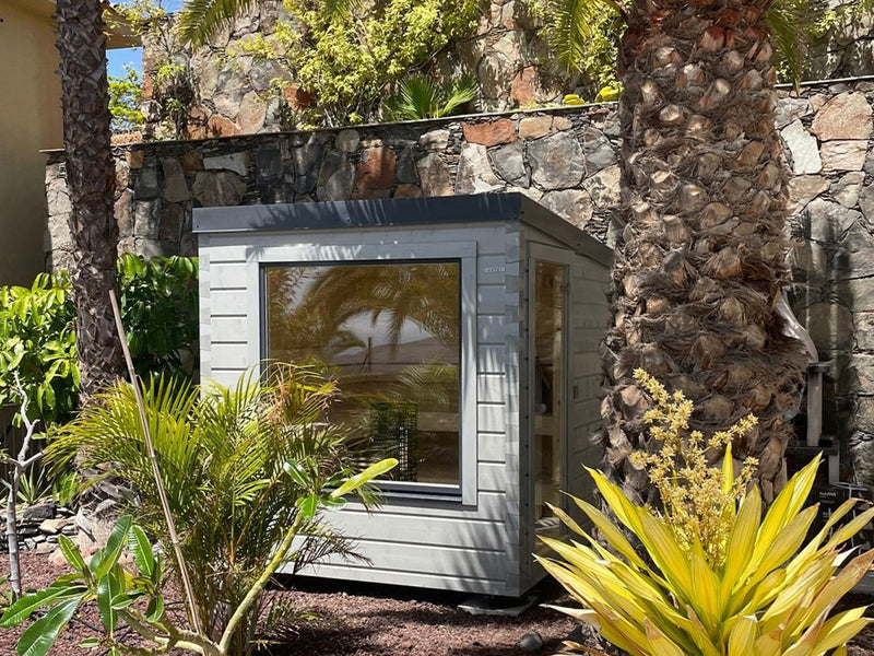 Graue Gartensauna mit großem Fenster zwischen Palmen und Natursteinmauer