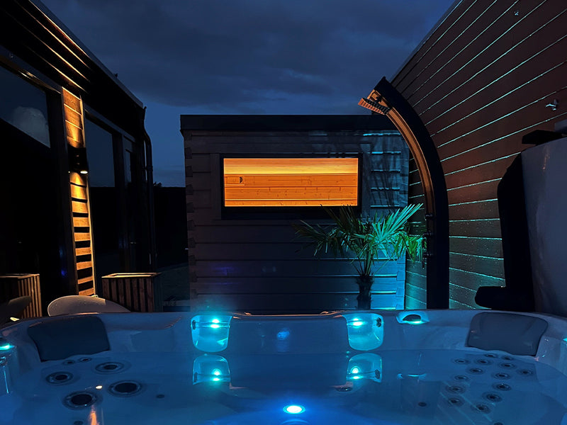 Beleuchtete dunkle Gartensauna hinter einem blau beleuchteten Whirlpool bei Nacht