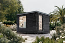 Dunkle Gartensauna Hetki Plus mit Fenster und Glastür zwischen Bäumen und Blüten