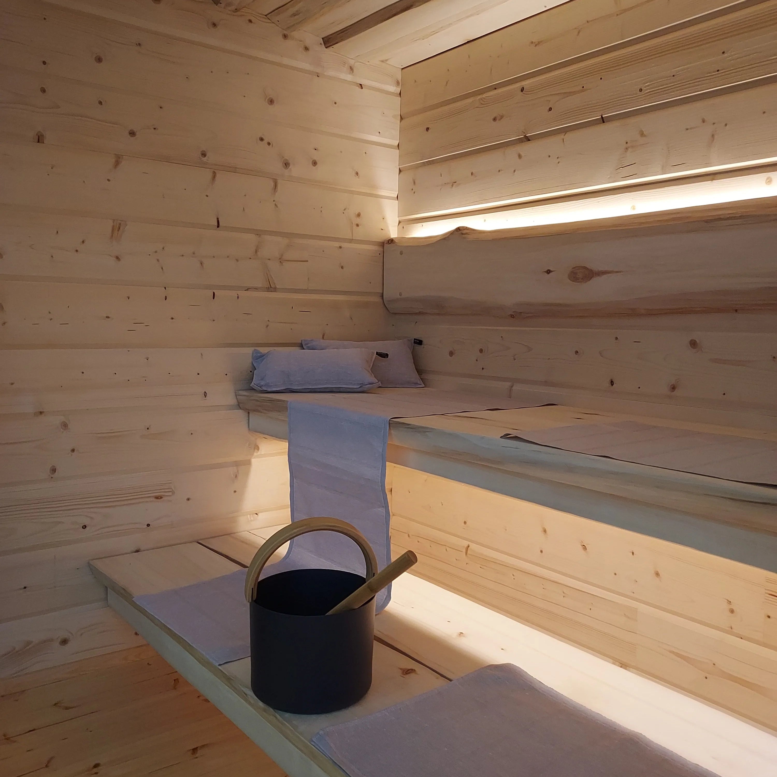 Beleuchteter Innenraum einer Gartensauna mit Holzbänken, Handtüchern und Aufgusseimer