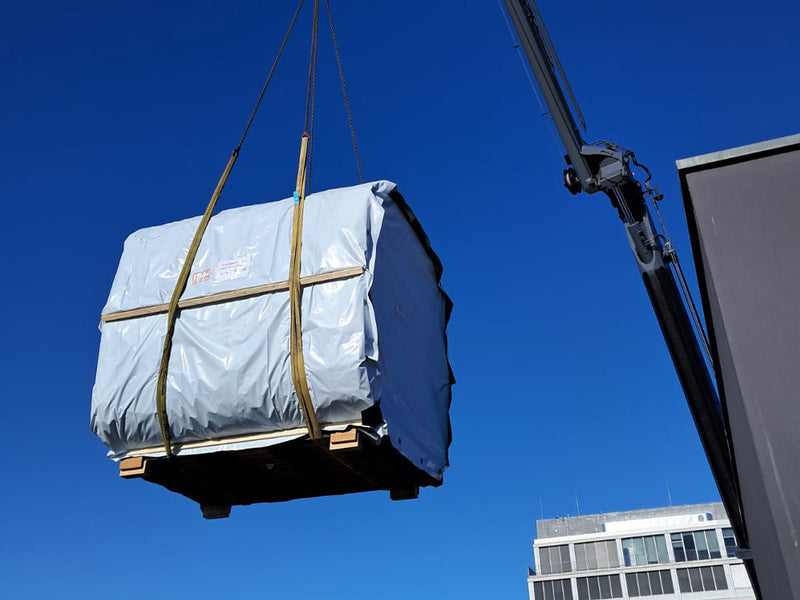 Großes verpacktes Saunamodul hängt an Hebegurten eines Krans vor einem Bürogebäude
