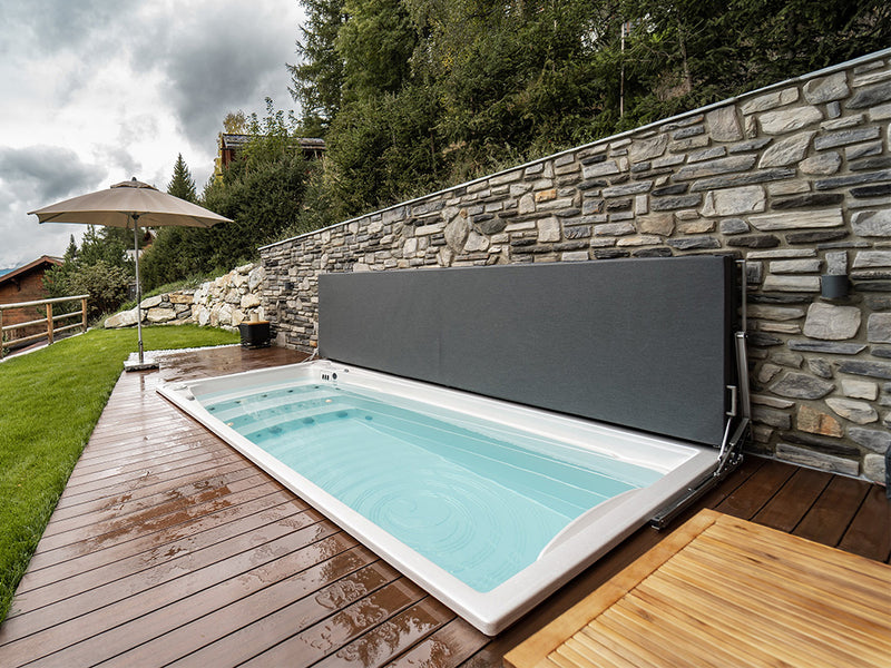 Swim-Spa mit geöffneter Abdeckung auf einer Holzterrasse vor einer Natursteinmauer