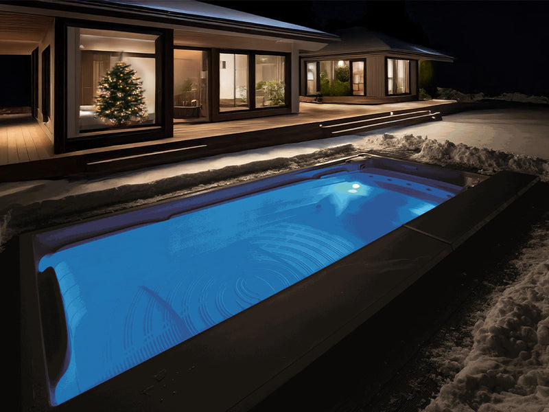 Blau beleuchteter Swim-Spa im Schnee vor einem Haus mit Weihnachtsbaum bei Nacht