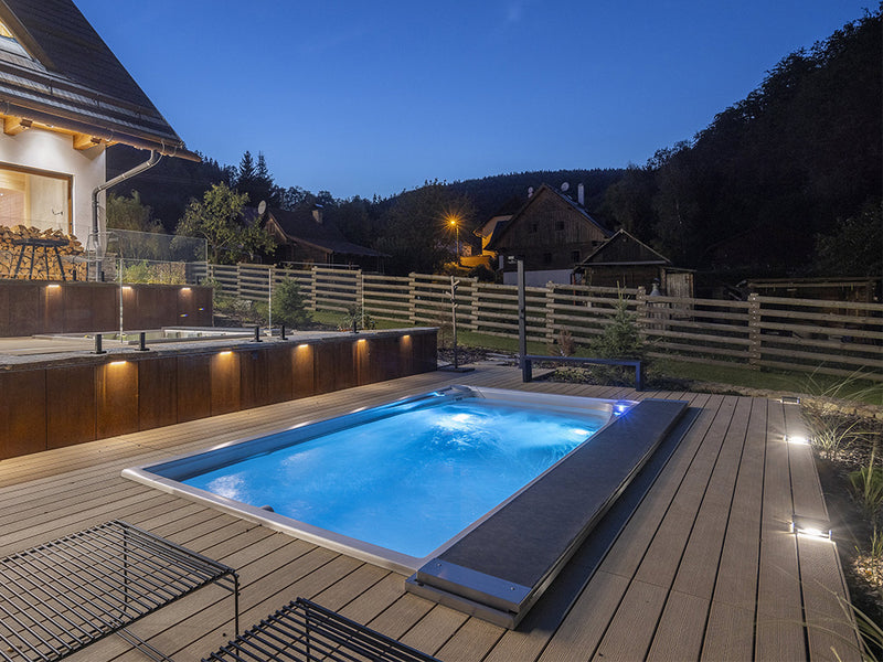 Blau beleuchteter, eingelassener Swim-Spa auf einer Holzterrasse bei Dämmerung