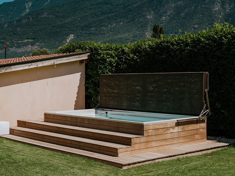 Swim-Spa mit Holzverkleidung, geöffneter Abdeckung und breiten Stufen im Garten vor Bergen