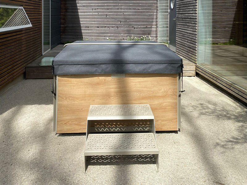 Geschlossener Whirlpool mit Holzverkleidung und Metalltreppe auf einer Terrasse zwischen Holzwänden