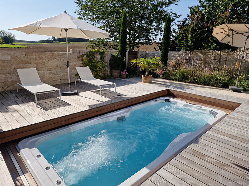 Sprudelnder VIVO SPA Waterfit Swim-Spa, eingelassen in eine Holzterrasse mit Liegen und Sonnenschirmen