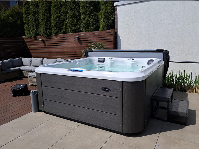 Geöffneter WELUXIA Whirlpool mit dunkler Verkleidung und Einstiegstreppe neben einer Gartenlounge