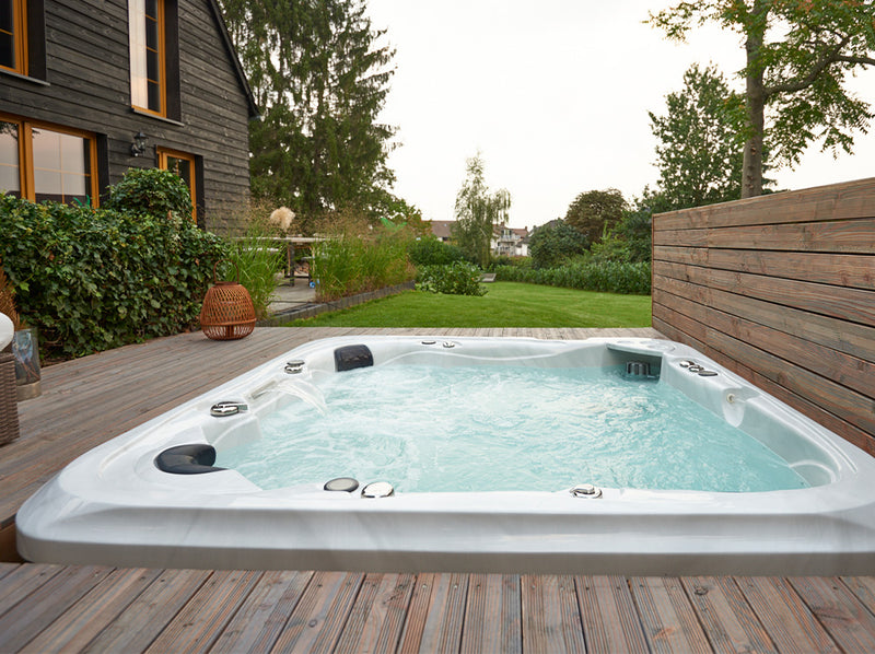 Sprudelnder WELUXIA Whirlpool, bodengleich in eine Holzterrasse neben einem dunklen Holzhaus eingelassen