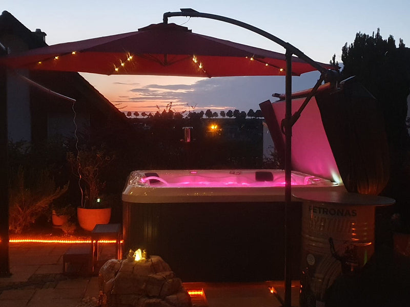 Pink beleuchteter WELUXIA Whirlpool unter einem roten Sonnenschirm mit Lichterkette bei Sonnenuntergang