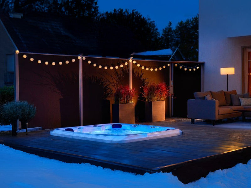 Blau beleuchteter, eingelassener WELUXIA Whirlpool auf einer verschneiten Terrasse mit Lichterkette