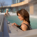 Frau bedient einen dampfenden Swim Spa im Schnee, während ein Mann im Wasser sitzt