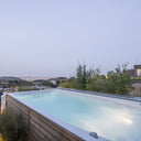Beleuchteter USSPA Swim Spa M mit Holzverkleidung auf einer Terrasse über der Stadt am Abend